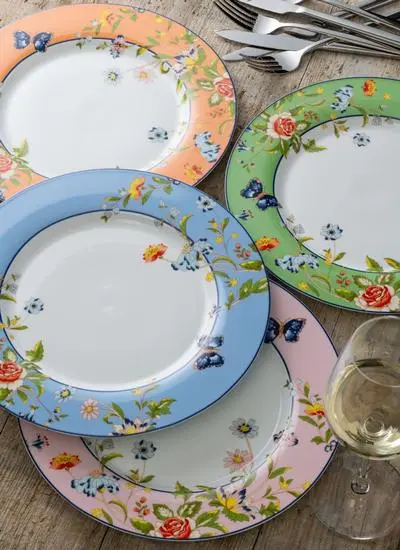 A variety of decorative floral plates and silverware on a wooden table, with a glass of white wine.